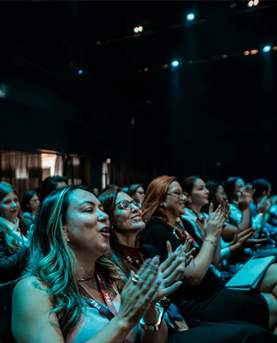 Participantes aplaudindo na plateia, iluminadas pela luz do palco